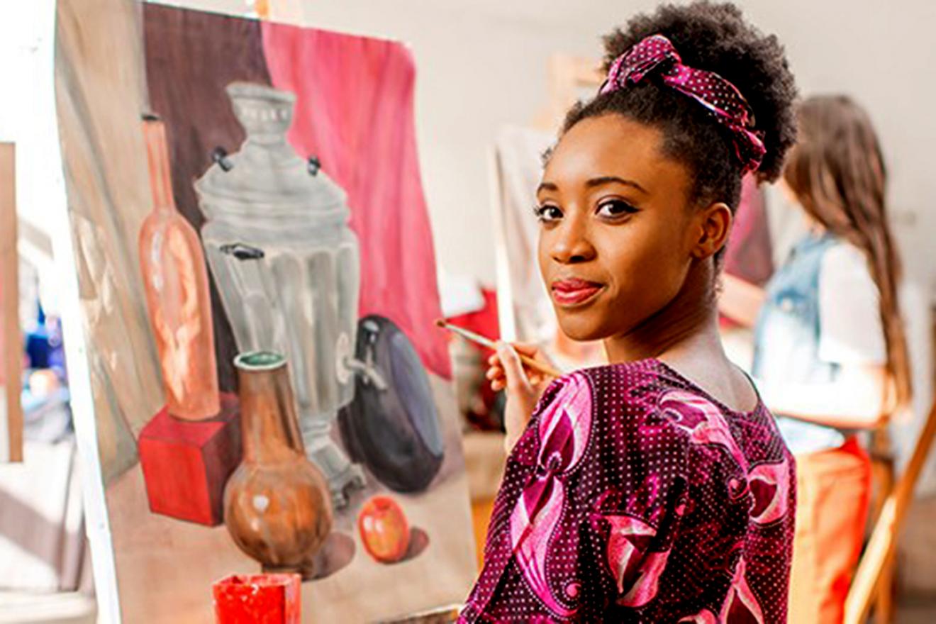 A woman poses in front of a painting in progress with a brush in hand.